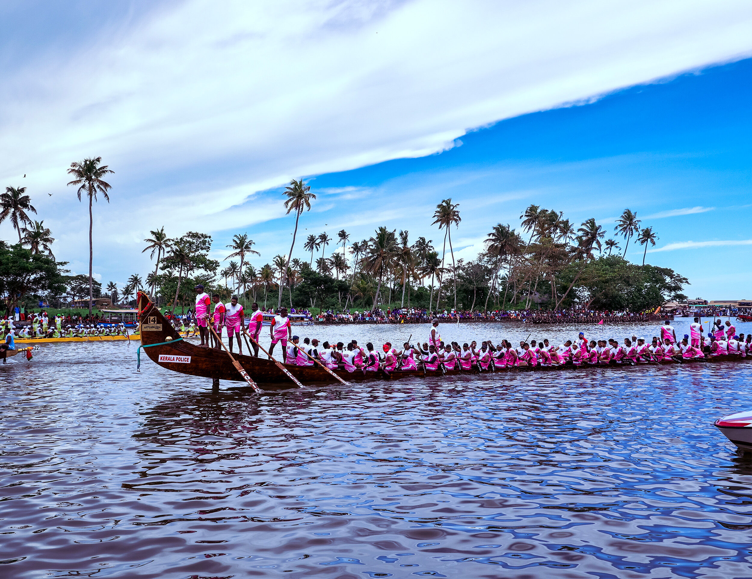Vibrant snake boat race in Kerala with rowers in pink uniforms paddling in sync during Onam Vallam Kali festival, surrounded by cheering crowds and palm-lined backwaters.